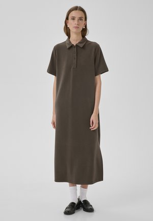 Ženska oblečena v rjavo polo obleko s kratkimi rokavi, bele nogavice, črne loaferje in biserne uhane, stoji ob enobarvnem sivi ozadju.