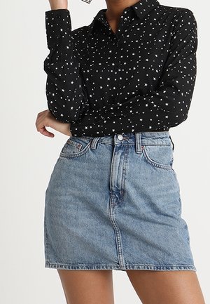 Kvinde iført en sort skjorte med lange ærmer og knapper, med et hvidt stjernemønster, samt en blå højttaljet denim nederdel, poserer med armene krydset.