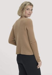 Blonde vrouw in een beige lange mouwen trui en zwarte broek, kijkt over haar schouder tegen een effen achtergrond.