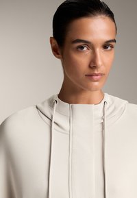 Hanorac bej deschis, realizat dintr-un material moale, având guler tip cowl, șnur de ajustare și un design minimalistic cu textură netedă.