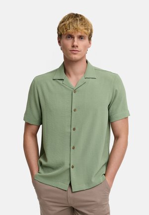 Homme blond aux cheveux courts portant une chemise vert clair à manches courtes et un pantalon beige, mains dans les poches, expression neutre.