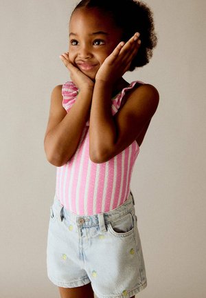 Jeune fille portant un haut rayé rose et un short en jean avec des ananas, souriante, les mains sur les joues, sur fond uni.