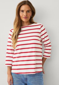 Top de rayas rojas y blancas de manga larga con escote barco y mangas 3/4, hecho de una suave tela de algodón. Combinado con jeans azules.
