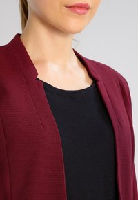 Blazer bordeaux avec une finition texturée, doté d'un col montant et d'un devant ouvert, superposé à un haut noir à col rond.