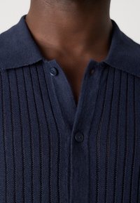Gros plan d'une personne à la peau foncée portant un polo en maille bleu marine avec une texture côtelée verticale et deux boutons visibles.