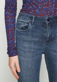 Vaqueros de mezclilla azul delgados con un acabado desvanecido. Presentan cinco bolsillos, un cierre de cremallera y un botón metálico oscuro. Textura suave y diseño elegante.