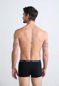 Schwarze Boxershorts für Männer mit einem gebrandeten Bündchen. Der Stoff wirkt glatt. Auffällige Tattoos sind am Rücken und am Arm sichtbar.