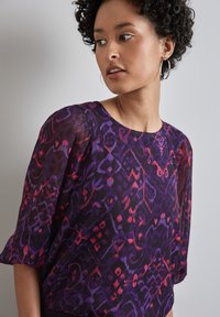 Blusa viola trasparente con un vivace motivo astratto in rosso e magenta. Presenta un collo a giro e maniche a sbuffo tre quarti.