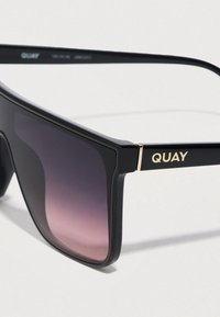 Μαύρα πλαστικά γυαλιά ηλίου με έντονο τετράγωνο σχέδιο, φακούς με διαβάθμιση από σκούρο σε ανοιχτό και χρυσό λογότυπο "QUAY" στον βραχίονα.