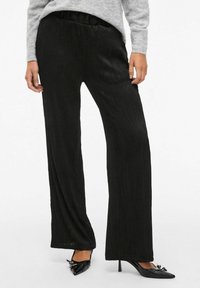 Pantalons larges noirs en tissu texturé avec des lignes verticales et une taille élastique, associés à des escarpins noirs pointus.