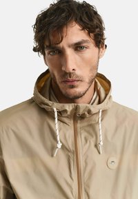 Giacca impermeabile beige con cappuccio, zip frontale e lacci bianchi con accenti rossi; badge con logo sul lato sinistro del petto.