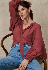 Blusa color ruggine con scollatura legata e maniche a palloncino, abbinata a jeans azzurri a vita alta. Il tessuto sembra leggero e morbido.