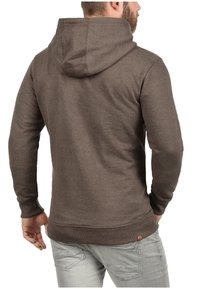 Sudadera marrón con capucha ajustable, puños y dobladillo acanalados, fabricada con un tejido suave y texturizado. Etiqueta visible cerca del dobladillo.