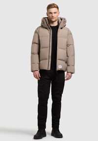 Beige donsjacket met een capuchon, voorzien van een gestikt patroon van horizontale secties. Zwarte accenten en een logopatch aan de zoom. Zwarte broek en laarzen.