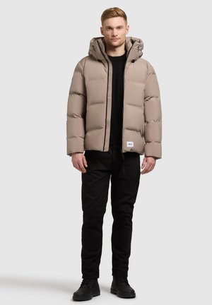 Beige donsjacket met een capuchon, voorzien van een gestikt patroon van horizontale secties. Zwarte accenten en een logopatch aan de zoom. Zwarte broek en laarzen.