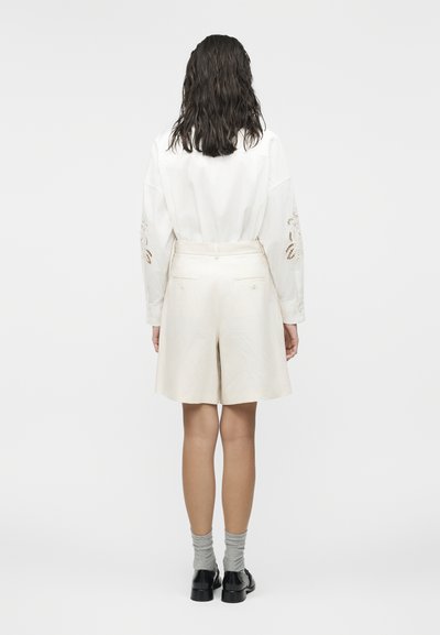 Femme aux cheveux foncés mi-longs portant une chemise blanche à manches longues avec des détails en dentelle, un short de couleur crème, des chaussettes grises et des chaussures noires, debout.