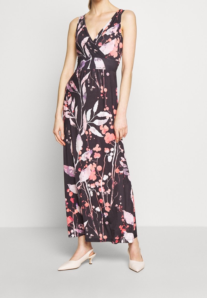 Femme portant une robe longue noire sans manches avec des motifs floraux roses et blancs et des chaussures beiges à talons bas pointues, debout sur un sol gris.