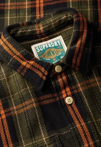 Flanellen shirt met een groen, marineblauw en oranje ruitpatroon. Heeft een knoopkraag, twee knopen en een merklabel van Superdry Workwear.