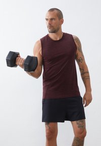 Μαρόν αμάνικο αθλητικό μπλουζάκι με υφή, σε συνδυασμό με μαύρες βερμούδες. Το άτομο κρατά έναν μαύρο εξάγωνο αλτήρα με το ένα χέρι.