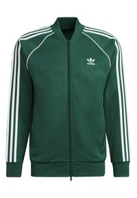 Grön zip-jacka i mjukt material med vit tre-stripes-design längs ärmarna och ribbade muddar och nederkant. Har Adidas-logotyp på bröstet.