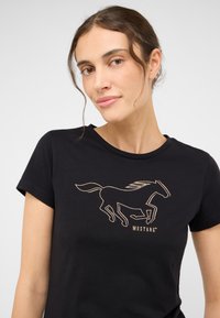 Čierne tričko vyrobené z mäkkej tkaniny, s kovovo zlatou grafikou bežiaceho koňa a textom "MUSTANG" nižšie. Krátke rukávy, okrúhly výstrih.
