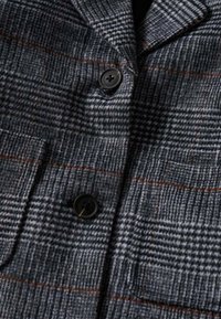 Blazer à carreaux gris en laine, avec un motif texturé et de fines rayures orange, comportant deux boutons noirs et une poche poitrine.