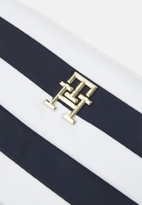 Přenosná taška pruhovaná v námořnické modři a bílé barvě s důrazem na zlatý geometrický logo akcent. Hladký povrch; zipové uzavírání nahoře.