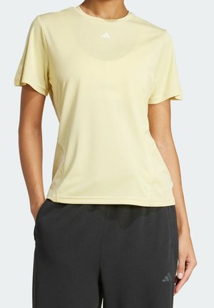 Personne portant un t-shirt de sport jaune pâle à manches courtes et un pantalon noir, debout les mains dans les poches devant un fond uni.