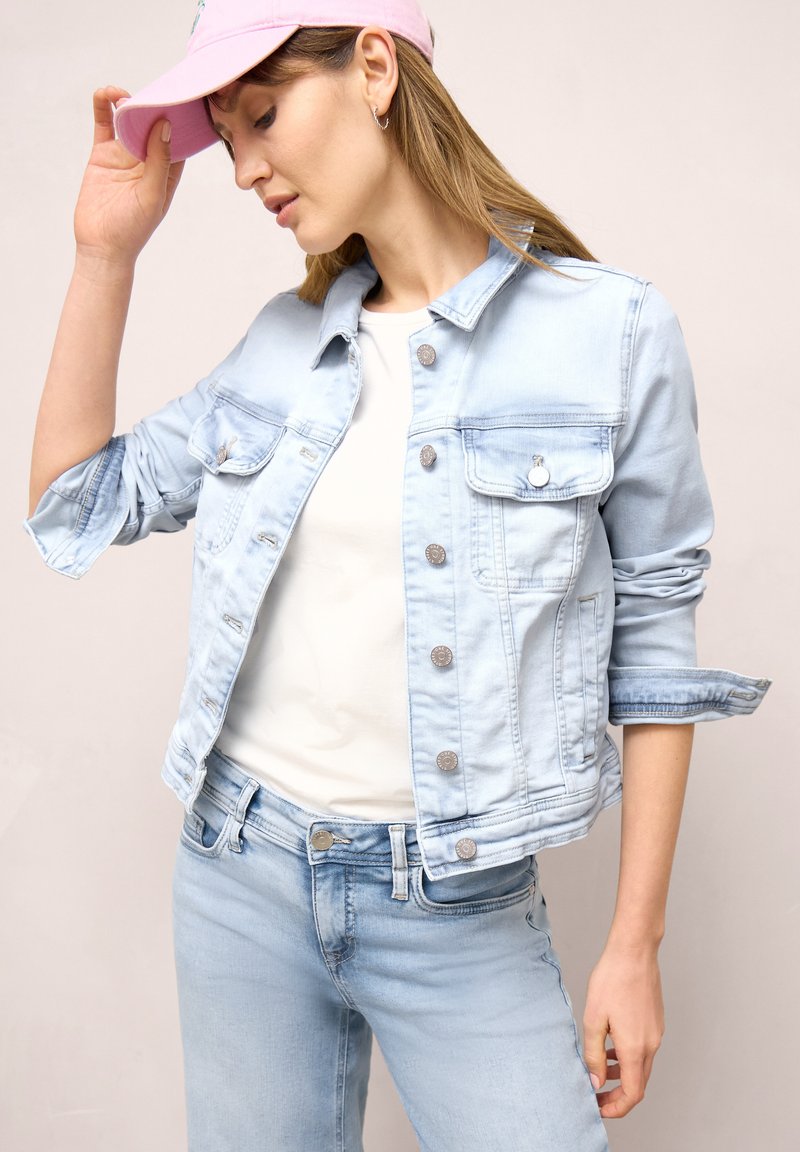 Kvinde iført lysblå cowboyjakke og jeans med hvid skjorte og lyserød kasket, hælder hovedet ned og rører ved skyggen på kasketten.