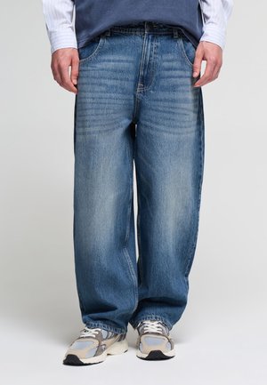 Persona che indossa jeans blu larghi e scarpe da ginnastica grigio-bianco, in piedi davanti a uno sfondo chiaro uniforme.
