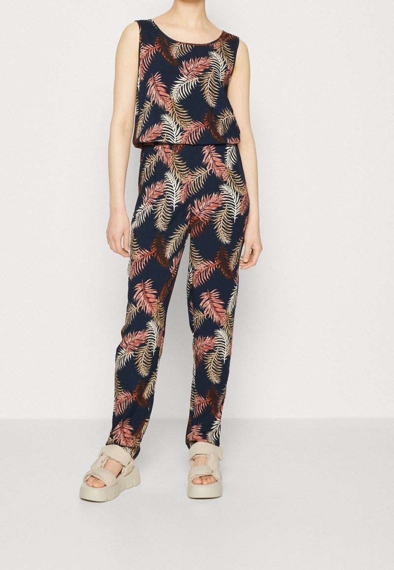 Femme portant une combinaison sans manches bleu marine avec des motifs de feuilles multicolores et des sandales à plateformes beige, debout devant un fond blanc uni.