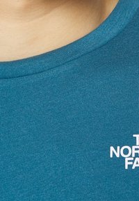 Teal bomullströja med rund hals och vit textlogotyp på framsidan. Slät textur, korta ärmar och inga ytterligare mönster.