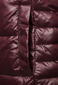 Bordeauxrote Steppjacke mit glänzender, gesteppter Textur, die horizontale Nähte und eine Reißverschlusstasche als Detail aufweist.