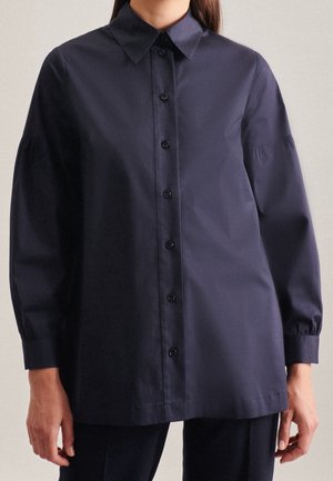 Mujer que lleva una camisa abotonada de manga larga de color azul marino oscuro con cuello puntiagudo y ajuste holgado.