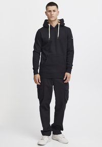 Sudadera negra con capucha y cordones, bolsillo frontal; combinada con pantalones cargo negros con bolsillos laterales y zapatillas blancas.