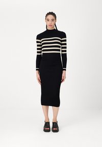 Črna pletena obleka z rebrastim vzorcem in smetanovimi vodoravnimi črtami, dolgo rokavi in prilegujočo silhueto, v kombinaciji z črnimi odprtimi sandalami.