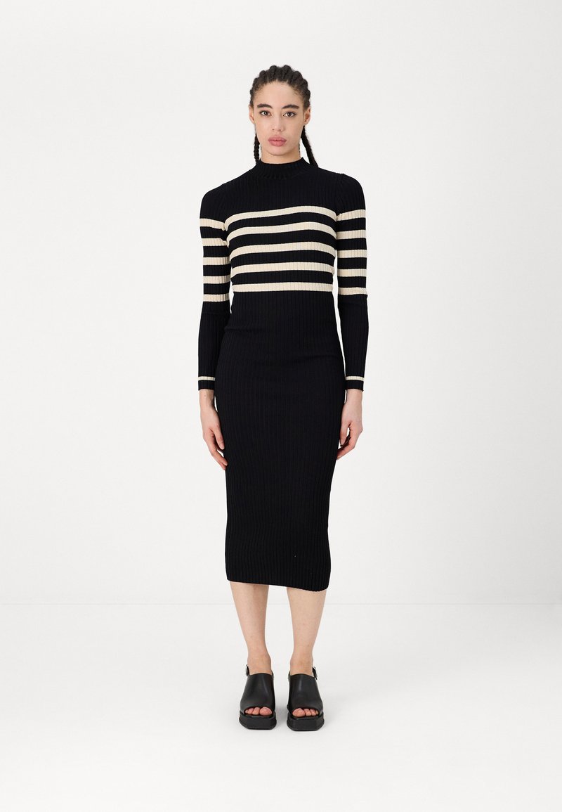 Črna pletena obleka z rebrastim vzorcem in smetanovimi vodoravnimi črtami, dolgo rokavi in prilegujočo silhueto, v kombinaciji z črnimi odprtimi sandalami.