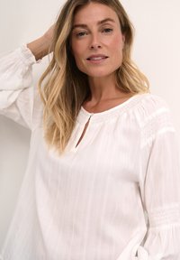 Vit blus med randigt mönster, med en nyckelhålsformad hals, rynkade ärmar och smockade sidodetaljer för textur och volym.