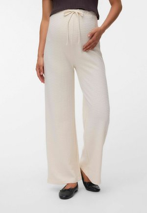 Femme portant un pantalon large texturé écru avec une taille à cordon et des ballerines noires, main posée sur la hanche.