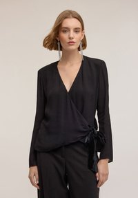 Blusa nera avvolgente con maniche lunghe, caratterizzata da un profondo scollo a V e un dettaglio annodato sul lato, realizzata in un tessuto morbido e leggero.