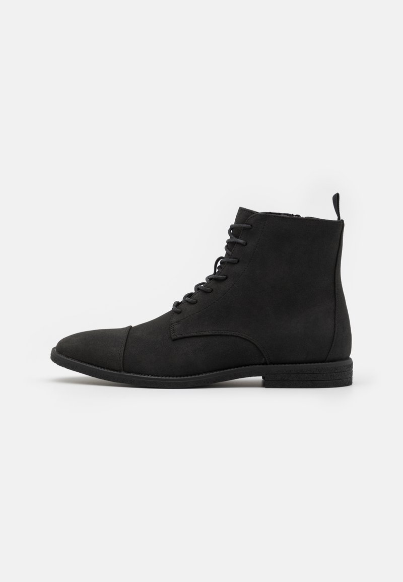 Bottines en daim noir avec bout arrondi, laçage à l'avant et semelle texturée discrète. Le design est minimaliste avec une languette à l'arrière.