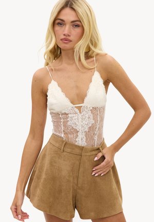 Femme blonde portant un top sans manches en dentelle blanche avec des bretelles fines et un short en daim beige taille haute, posant avec une main sur la hanche.