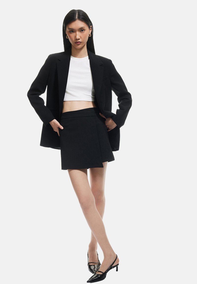 Blazer negro ajustado sobre un top corto blanco, combinado con una falda envolvente negra y tacones negros. Textura suave, diseño moderno, estilo minimalista.