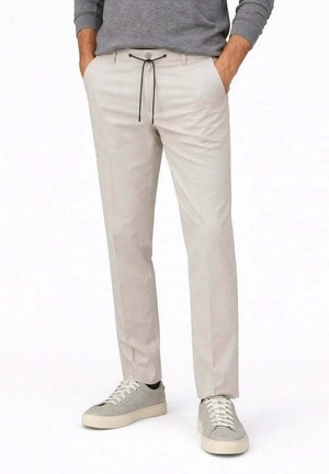 Uomo che indossa pantaloni beige aderenti con coulisse nera, maglia grigia a maniche lunghe e sneakers grigio chiaro, con le mani in tasca.