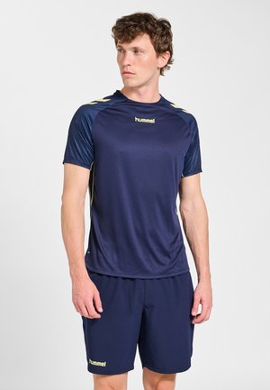 Man draagt een navyblauw hummel sportshirt en korte broek, kijkt opzij, staat tegen een effen witte achtergrond.