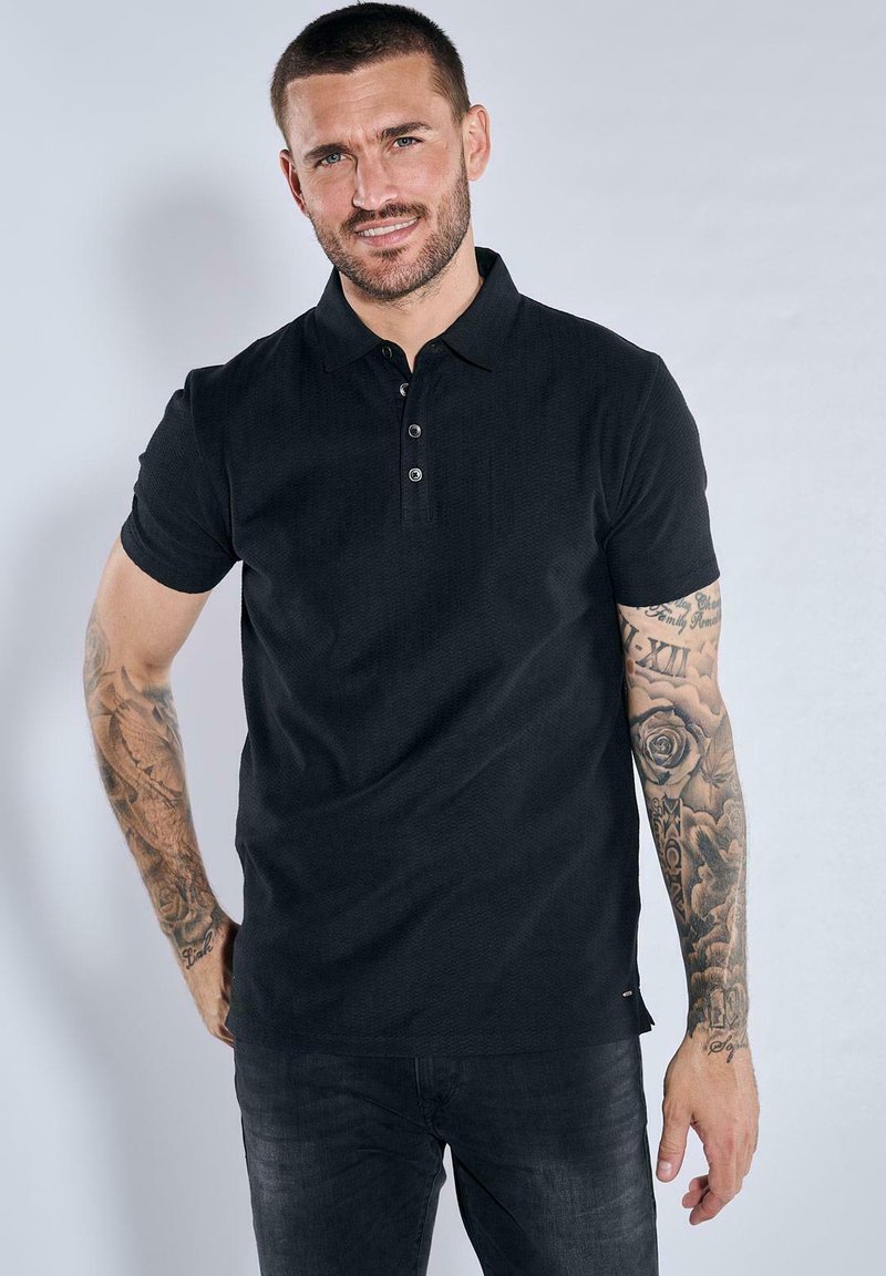 Black polo shirt with short sleeves, textured fabric, three-button placket, and a fitted design. Worn with dark jeans, accentuated tattoos visible.