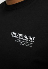 Schwarzes Baumwoll-T-Shirt mit weißem Aufdruck, der "THE DRYMART MEMBERS ONLY HOTEL & BAR" liest, einschließlich einer dekorativen Schrift darunter.