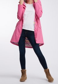 Donna con un impermeabile rosa sopra una camicia bianca, jeans skinny scuri e stivali beige con lacci, in piedi con le gambe divaricate su uno sfondo neutro.