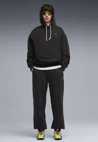 Felpa nera con cappuccio e lacci bianchi, abbinata a pantaloni neri larghi. Occhiali da sole con tonalità verde e sneakers dai colori vivaci completano l'outfit.