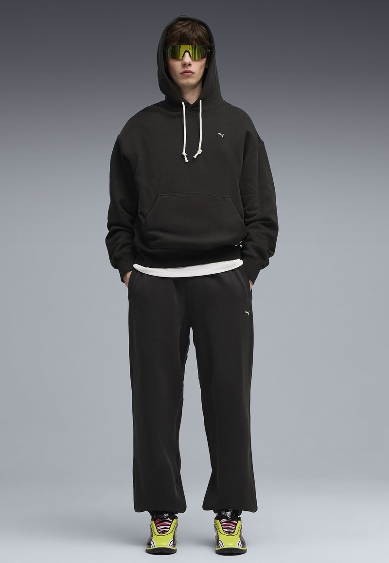 Felpa nera con cappuccio e lacci bianchi, abbinata a pantaloni neri larghi. Occhiali da sole con tonalità verde e sneakers dai colori vivaci completano l'outfit.